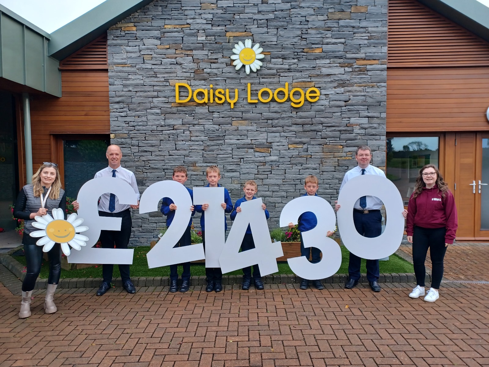 A group of BB Leaders and young members in front of Cancer Fund for Children, Daisy Lodge, holding cut out figures to represent £21430 raised.