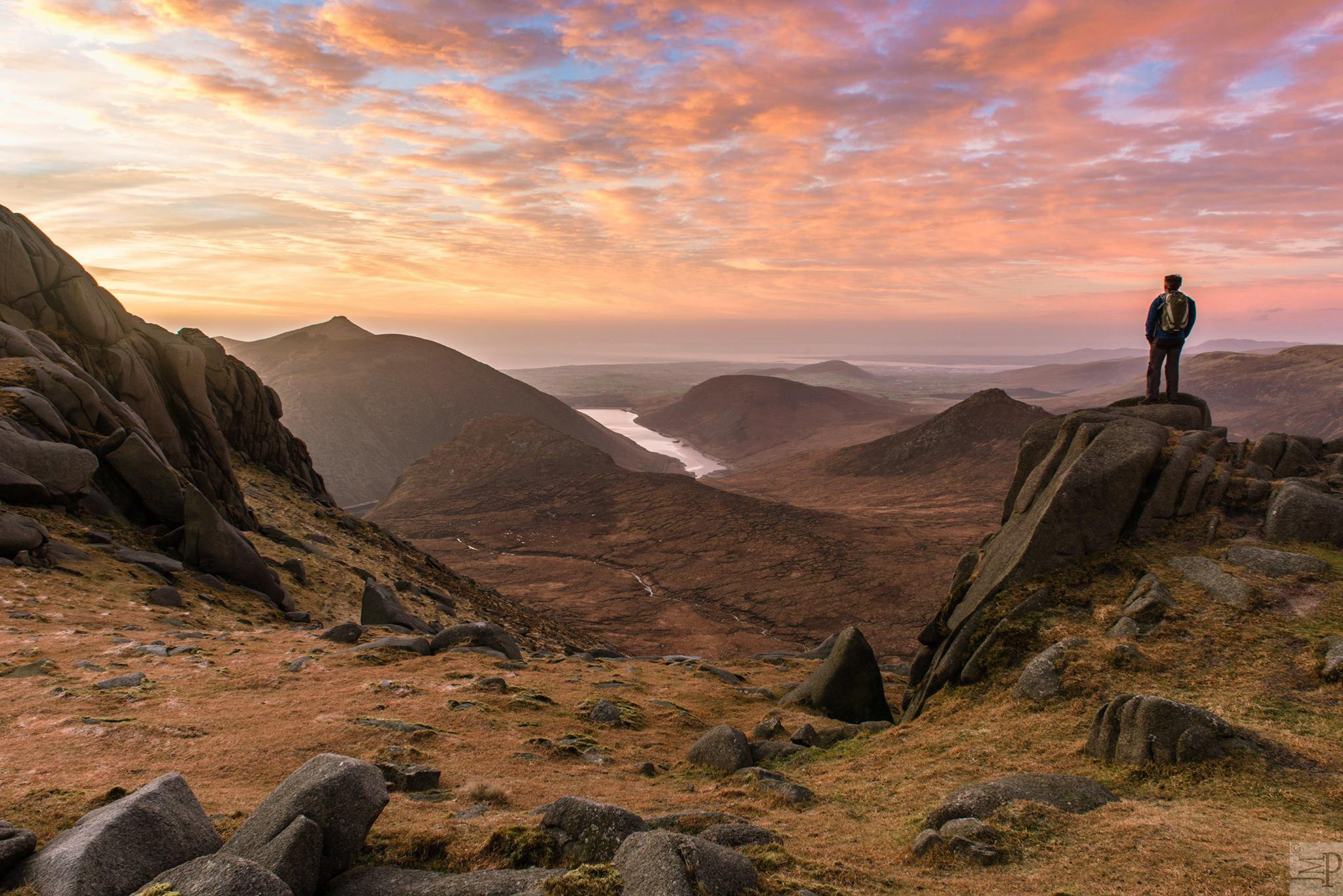 a man standing on a rock in the mourne mountains, looking out at a sunset over spelga dam.